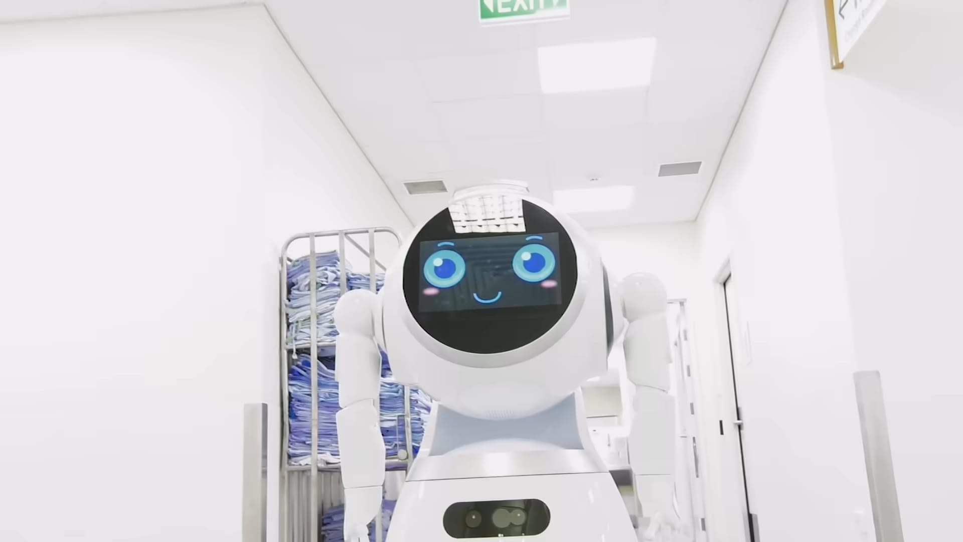 Un robot humanoide con una pantalla en lugar de rostro avanza por un pasillo hospitalario junto a carros de ropa de cama.
