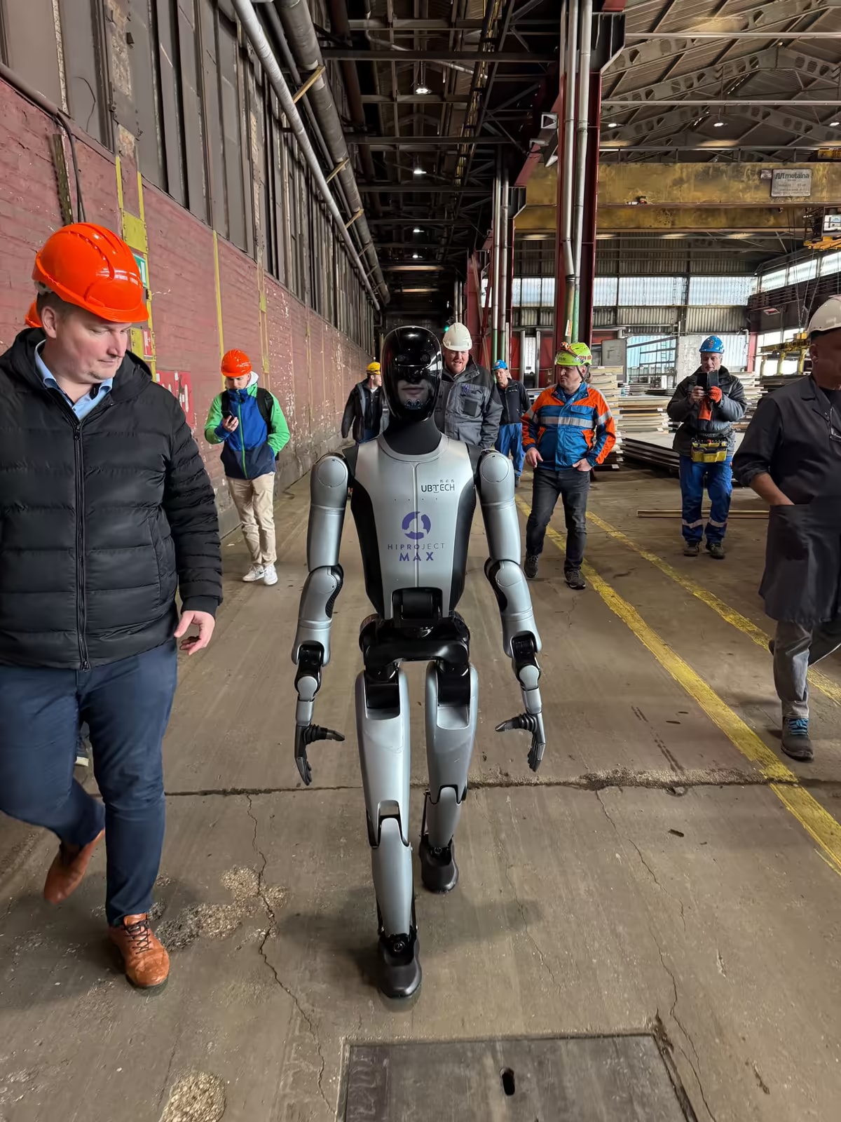 MAX geht durch eine Industriehalle zwischen Arbeitern mit Schutzhelmen.