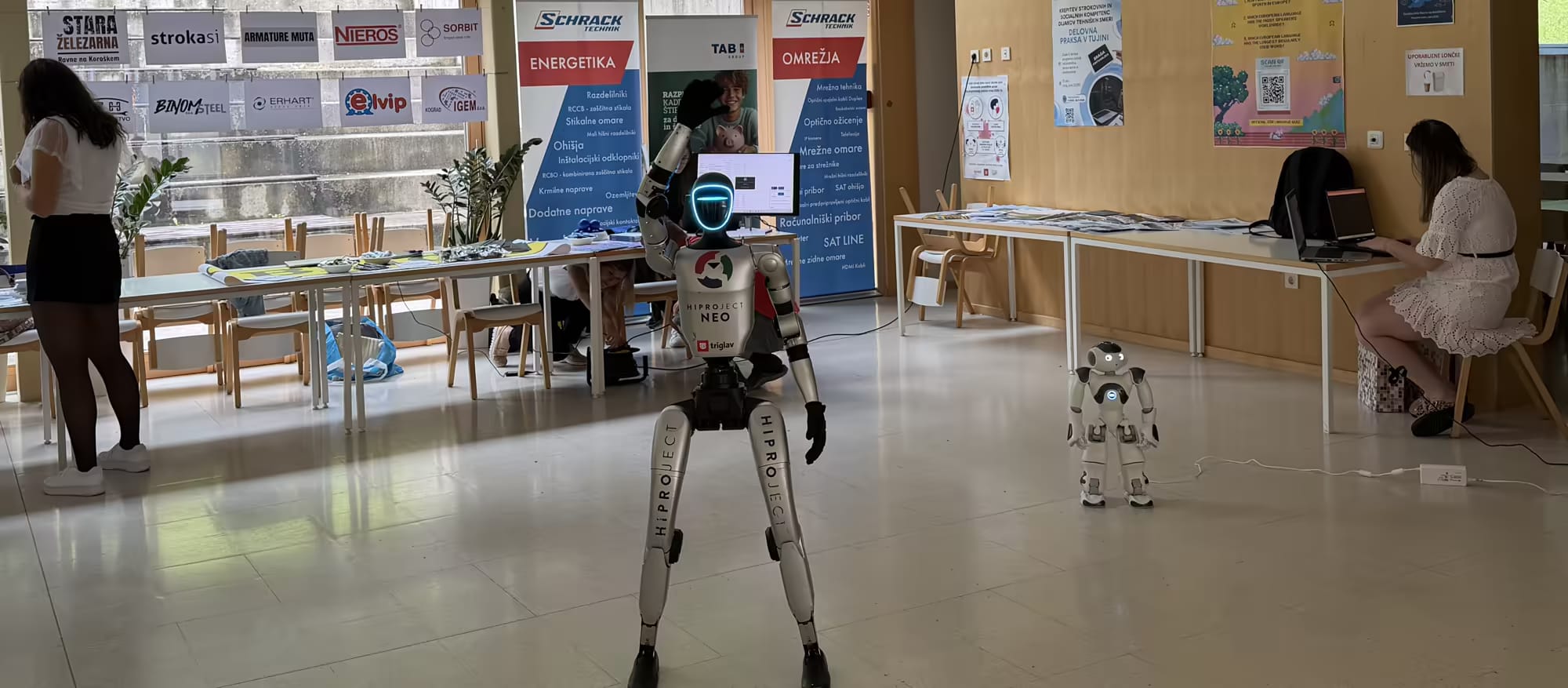 Robot NEO z dvignjeno roko v šolski avli med stojnicami, ob njem manjši humanoidni robot, dijaki delajo za mizami