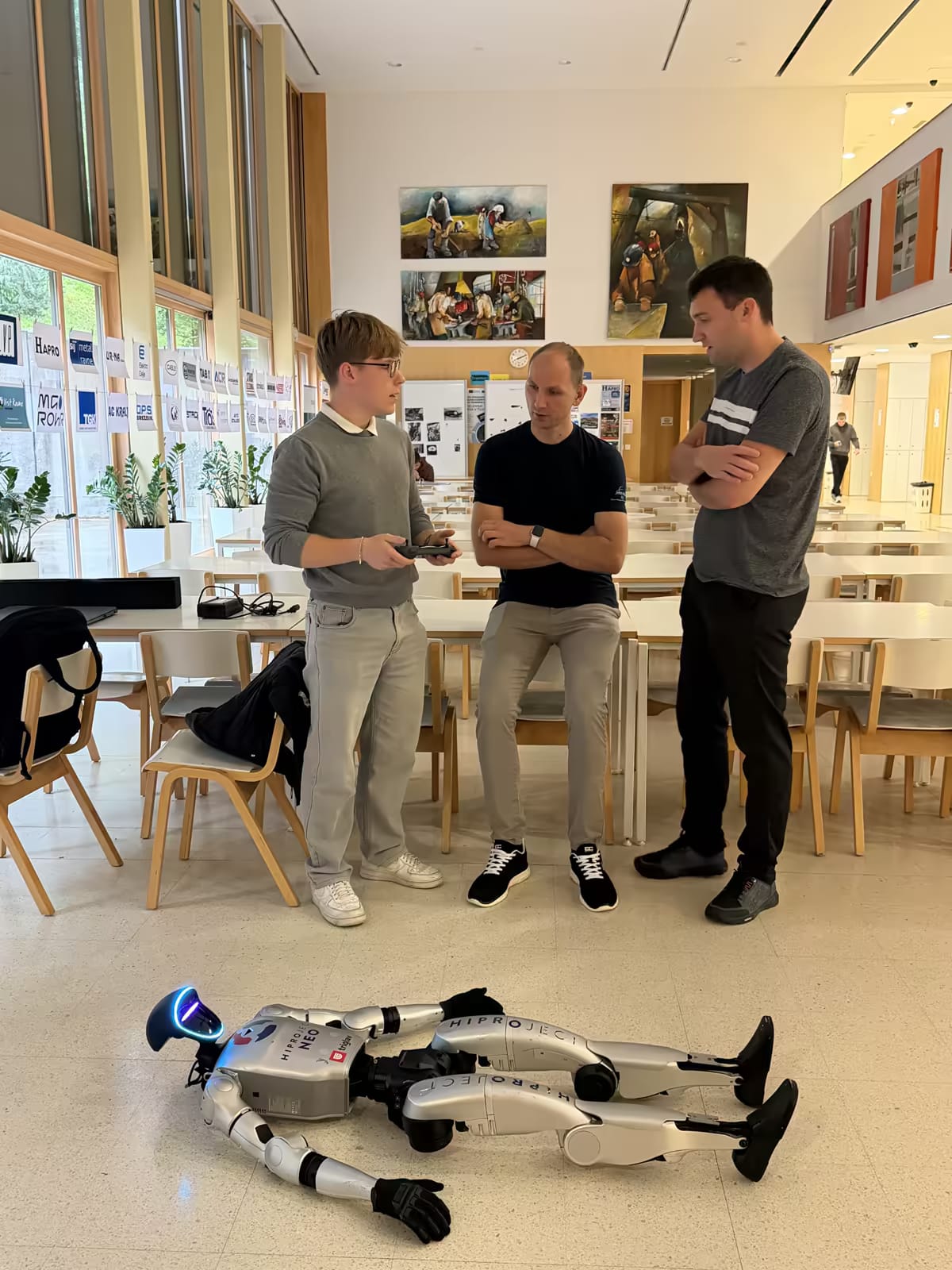 Robot NEO leži na tlaku šolske avle, dijak ga upravlja z ročnim krmilnikom, ob njem dva obiskovalca